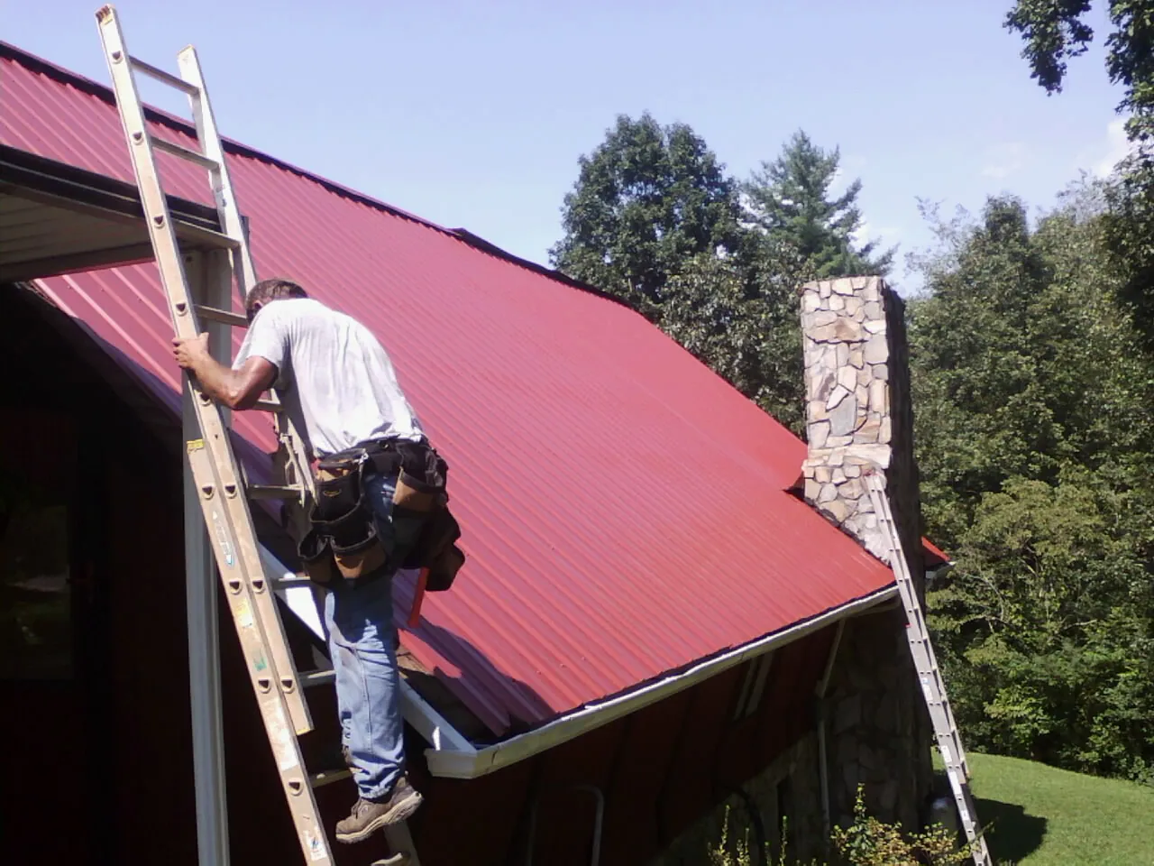 Roofer on a ladder beside a red metal roof and stone chimney — Soffit & Fascia Repair by Roofing Brothers in Edmond
